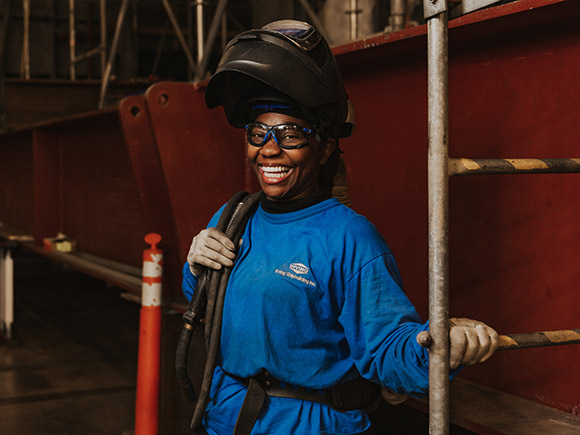 A welder stands in an industrial setting. She wears a long-sleeved shirt, safety goggles, gloves and a welding helmet.