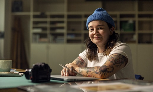 Anisa, a Graphic Design graduate, sits at a table and smiles while her hands rest on a closed laptop.