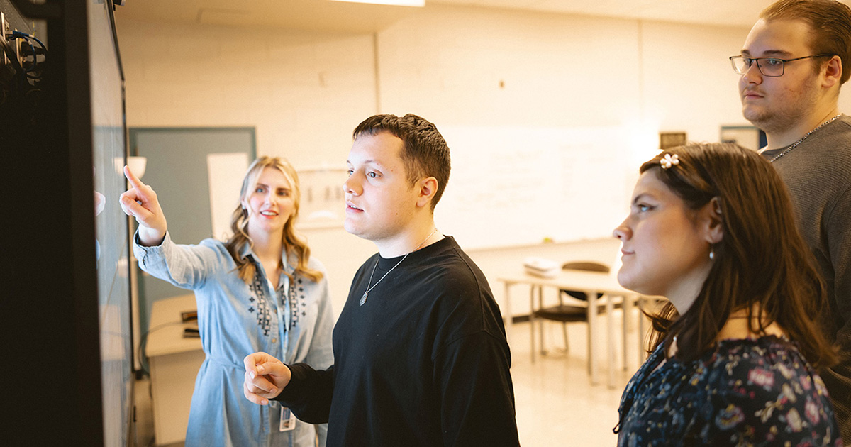 A group of students stand around a chalkboard while the teacher points out information. 