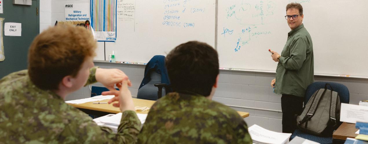Students in military-style uniforms listening to an instructor writing on a whiteboard.