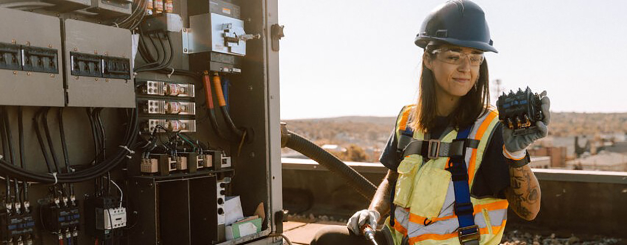 Woman wearing safety gear works on a heat pump on a roof top.