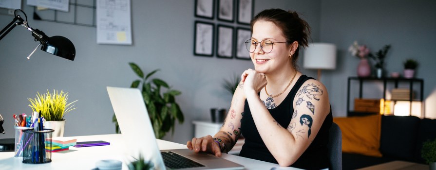 A smiling woman with tattoos, wearing glasses, looking at her laptop in her living room.