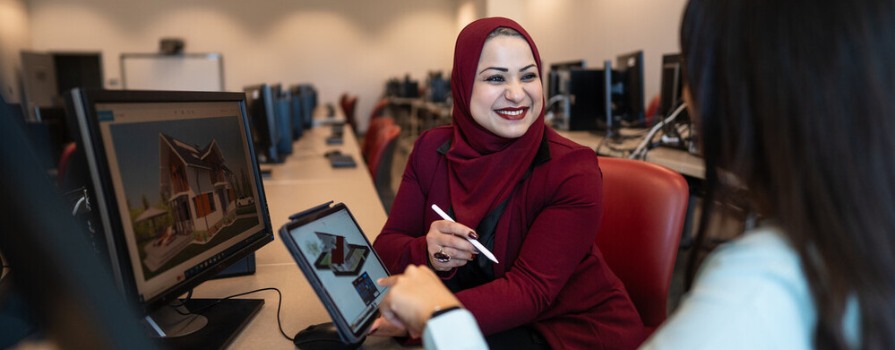 A woman wearing a hijab in a computer lab engages in a friendly conversation with another woman as they both examine designs on a tablet.