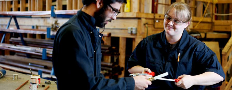 A man and a woman, dressed in matching dark blue work attire, converse while holding pieces of plumbing equipment in a workshop.