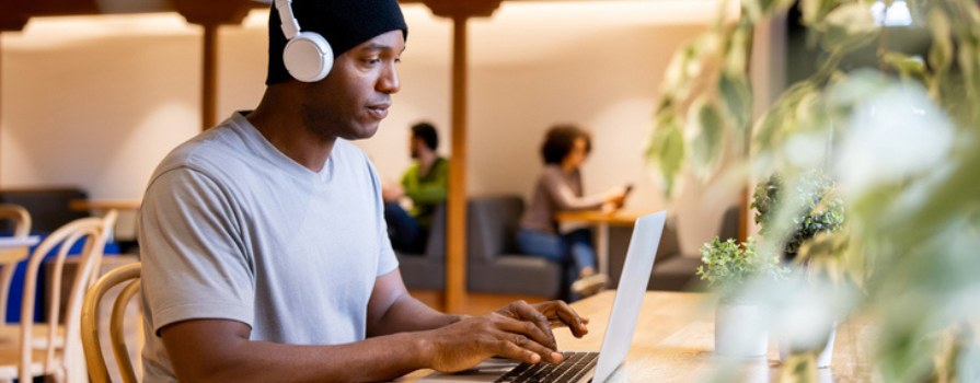 A focused man wearing headphones working on a laptop in a brightly lit, public space.