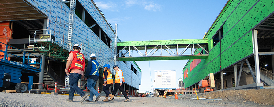 Four construction workers wearing hard hats, reflective vests and boots walk site-by-side through a construction site. There are buildings under construction in the background.