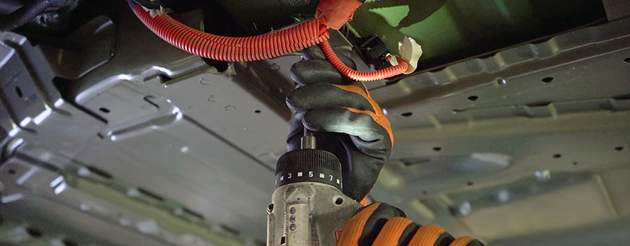 A close-up photo of hands in heavy-duty gloves using specialized equipment to work on the underside of an electric vehicle.