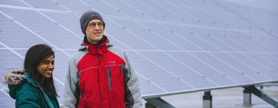 A man and woman smiling wearing winter attire stand side by side in front of a large solar panel installation.