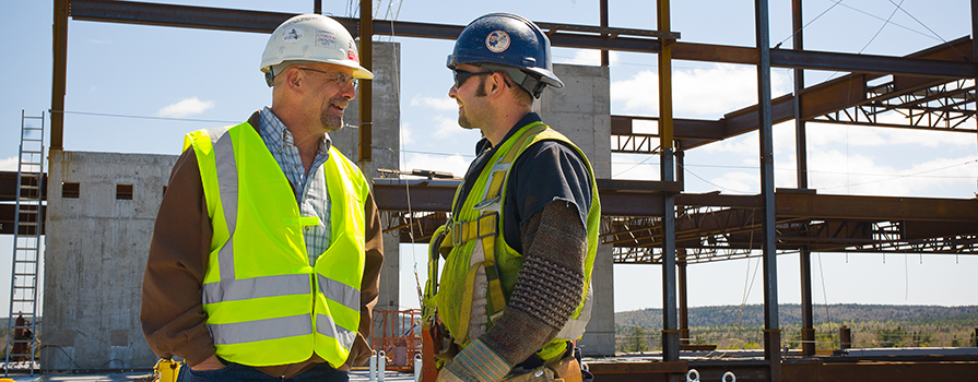 Two construction workers in reflective vests and hard hats speak with each other at a construction hob site. Both are smiling and facing one another in conversation.