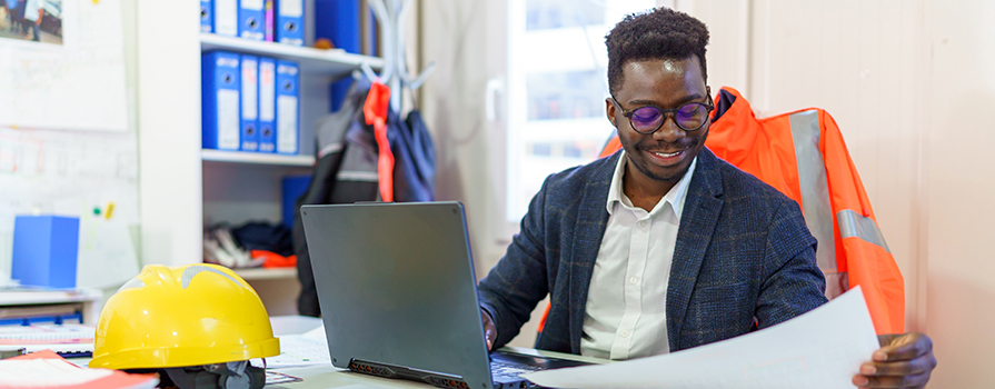 An office worker sits at a desk in front of a laptop computer. There are papers and a hard hat on the desk, and shelves with binders in the background. He looks at paperwork in his left hand and smiles.