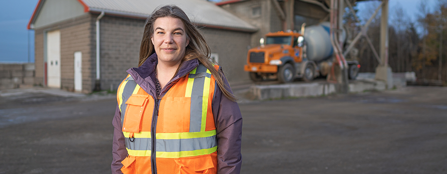 A construction worker wearing a warm coat and reflective vest stands at a construction site with a brick building and concrete truck in the background. She smiles slightly while facing the camera with the wind blowing her long hair away from her face.
