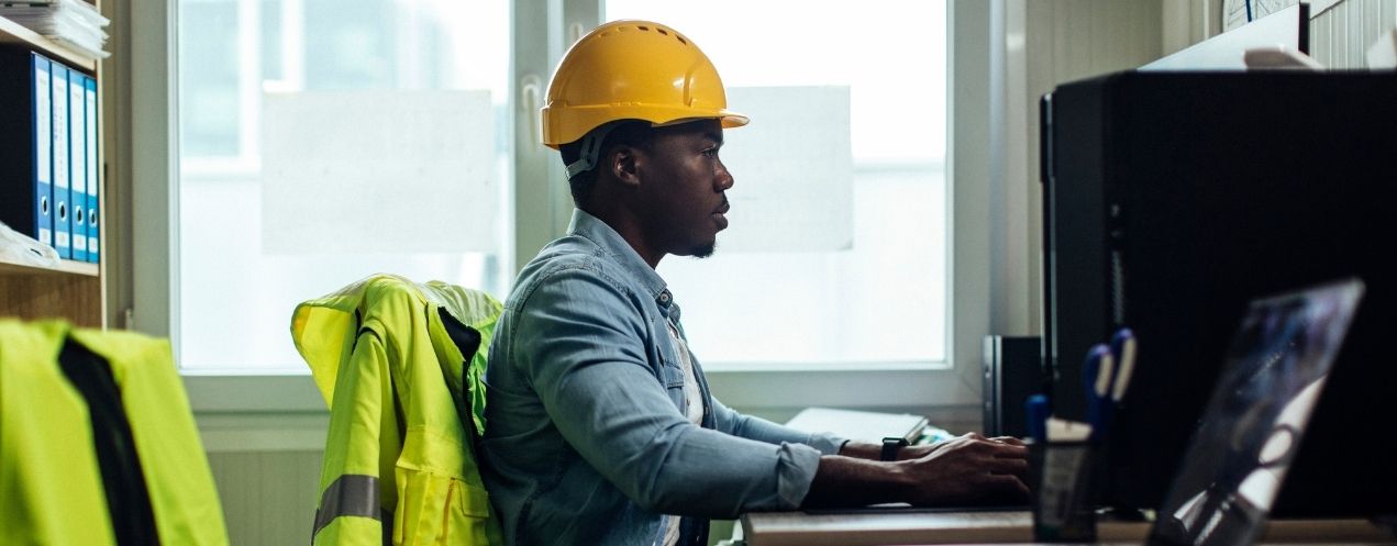 A maintenance professional wearing a hard hat using a computer in an industrial work environment.