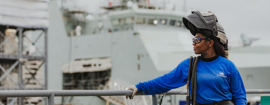 Woman with welding equipment looks out over the rail of a ship.