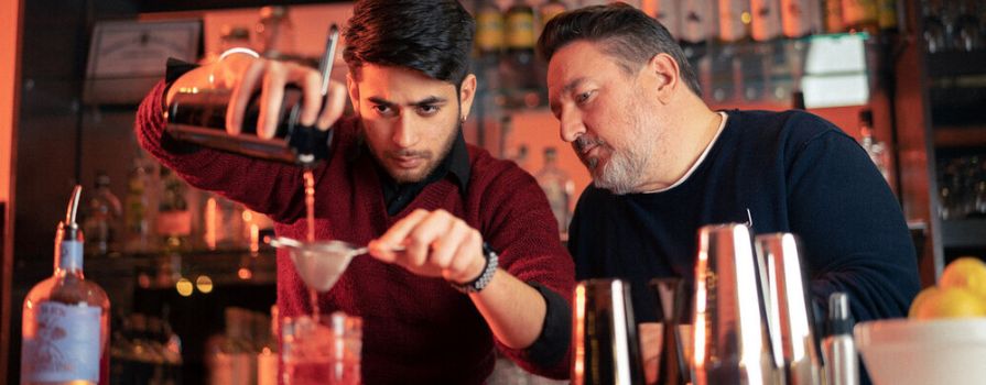 A supportive male instructor attentively observes as a male student skillfully pours a beverage through a strainer into a glass in a bar environment.