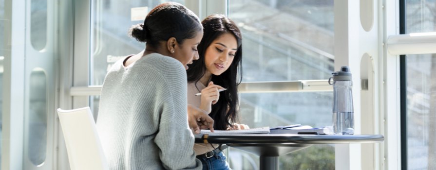 Two women sit at a table beside a large window and look a papers spread out on a table.