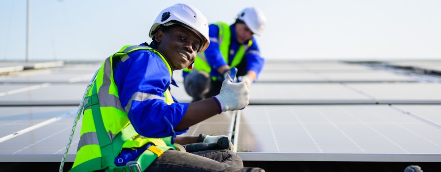 Two men wearing brightly coloured safety vest, hard hats and fall protection harnesses are working on solar panels, one giving a thumbs up, the other focused.