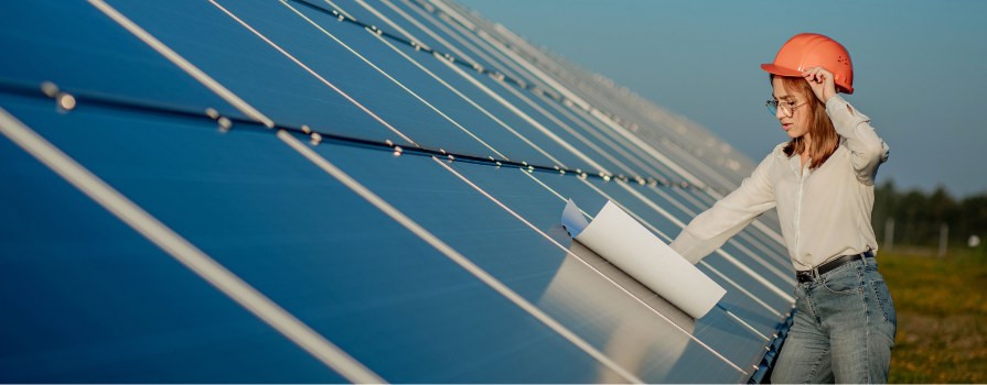 A woman in an orange hard hat leans against a solar panel, studying the contents of a large printout.