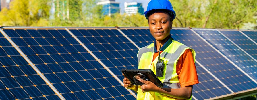 A woman wearing a bright safety vest and a blue hard hat is standing in front of a large solar panel holding a tablet.