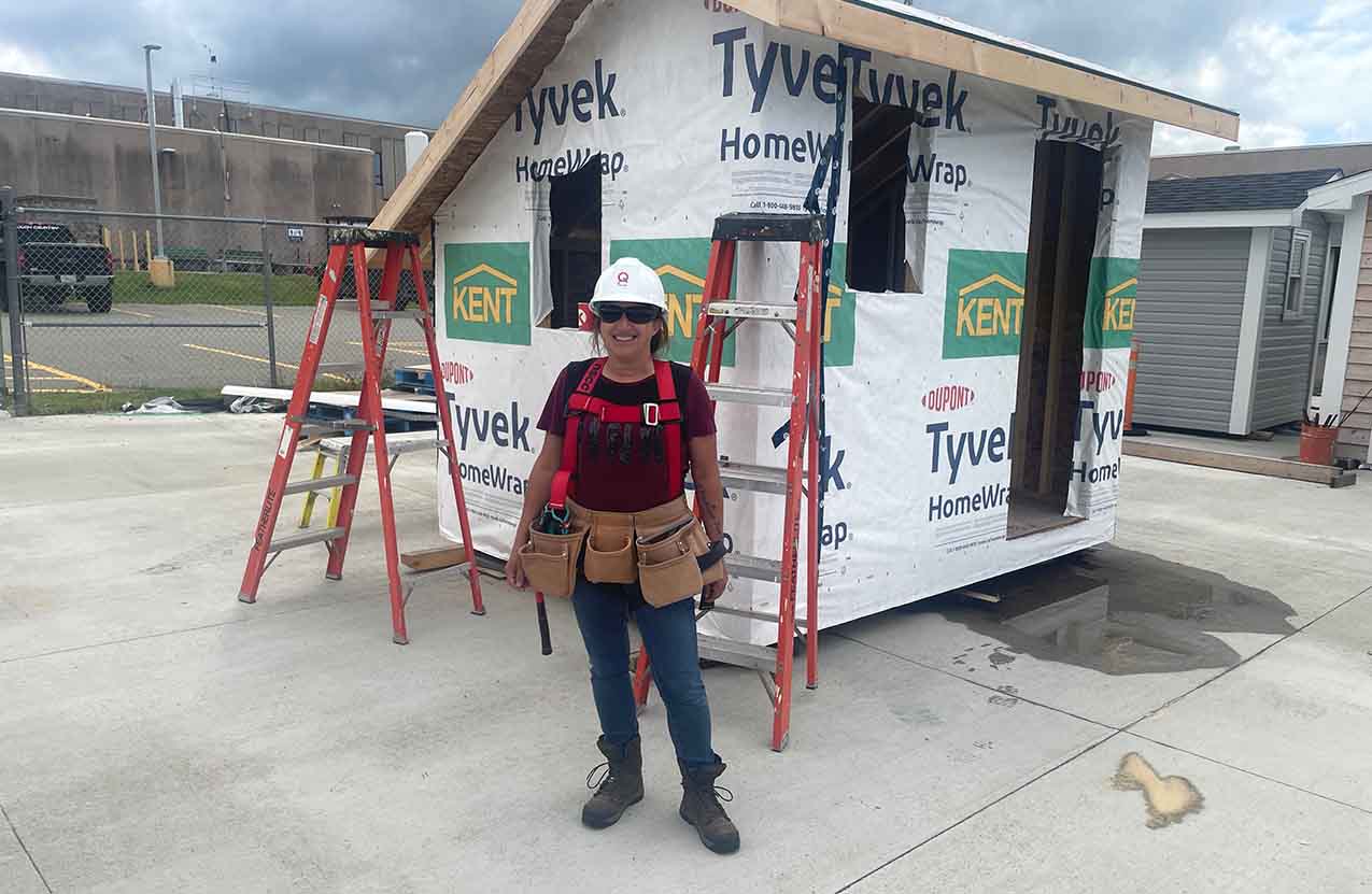 A cheerful woman, wearing a hard hat and professional trade attire, stands proudly in front of an under-construction shed.