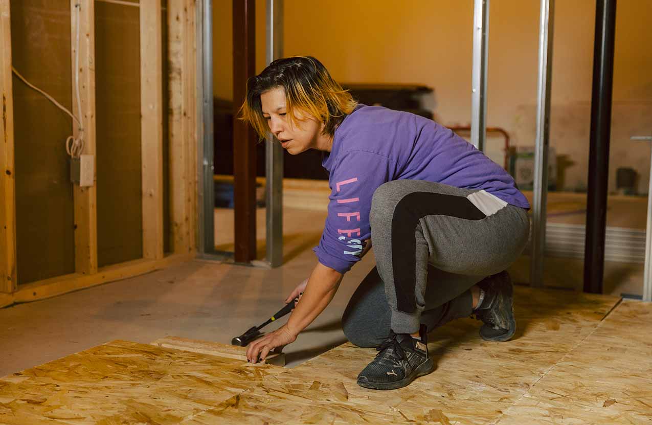 Woman working in a wooden shed frame kneeling over holding a hammer in her hand.