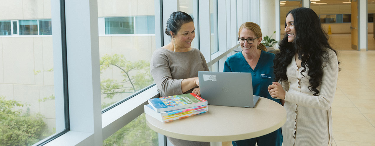 Three students stand at a round table next to tall windows in a campus hallway.