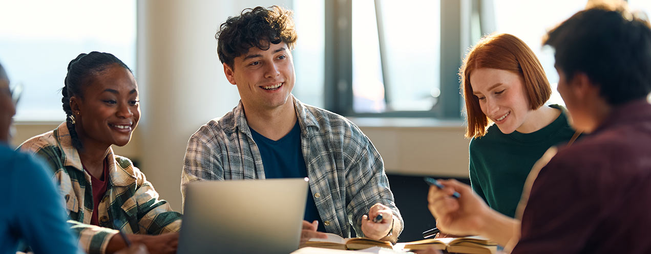 A group of students sit around a table and chat in a classroom with natural light.
