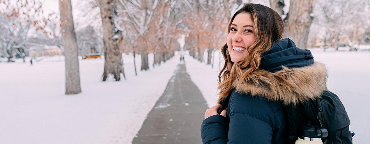 A student in a winter coat carrying a backpack looks back over her left shoulder and smiles at the camera in a wintery outdoor scene.