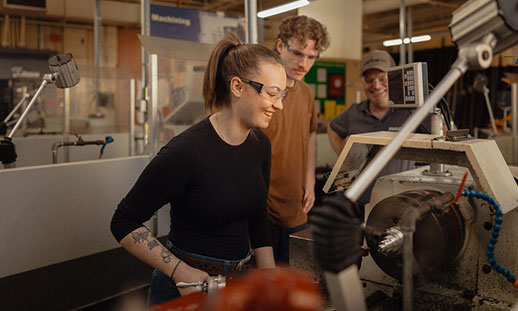 Three people stand in an NSCC Precision Machining workshop. One uses a machine while the other two watch.