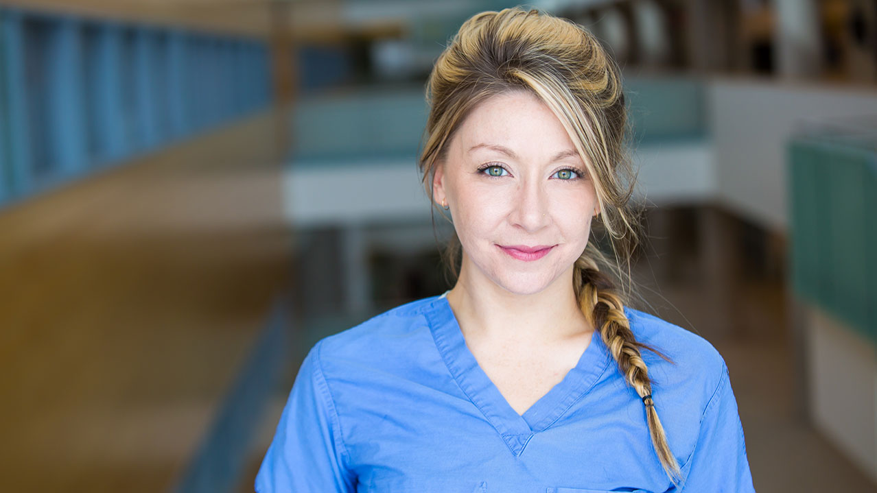 A woman in a blue shirt with a braid in her hair looks straight ahead and smiles against an out-of-focus background.