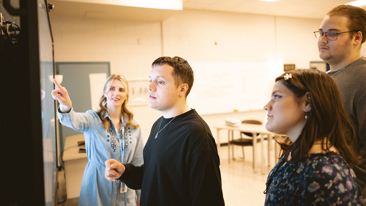 Three students stand facing a whiteboard while a smiling instructor points at the whiteboard.