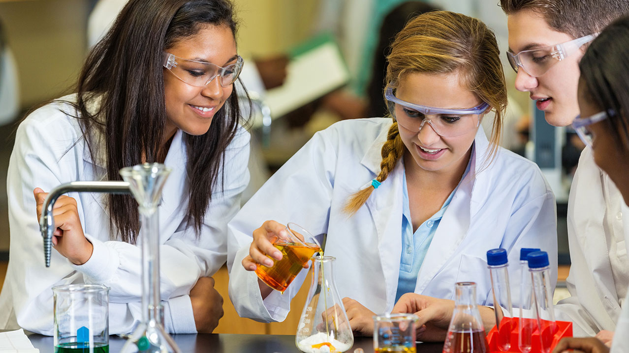 Four students, wearing white lab coats and safety glasses, conduct an experiment in a science lab.