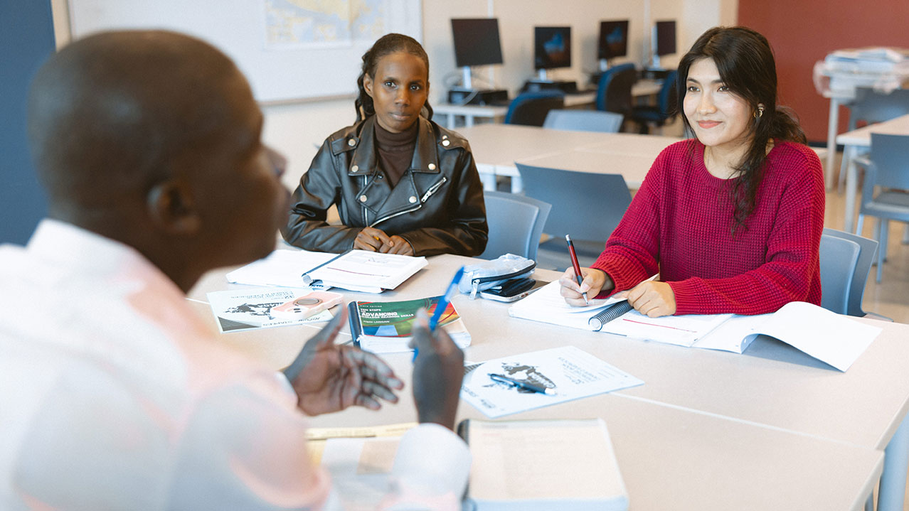 Three students sit together discussing something at a table covered in books and notes in a classroom setting. 
