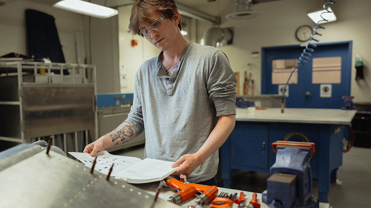A student wearing safety glasses in a workshop is seen reading a manual.