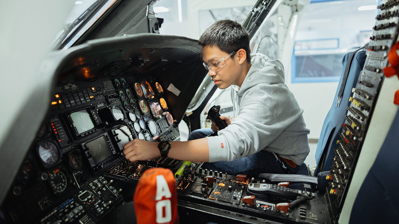 A student wearing safety glasses works on the inside electronics panel of an aircraft.