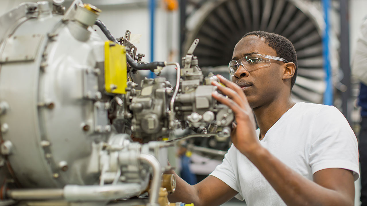 A student wearing safety goggles works on the inside of a plane's side engine.