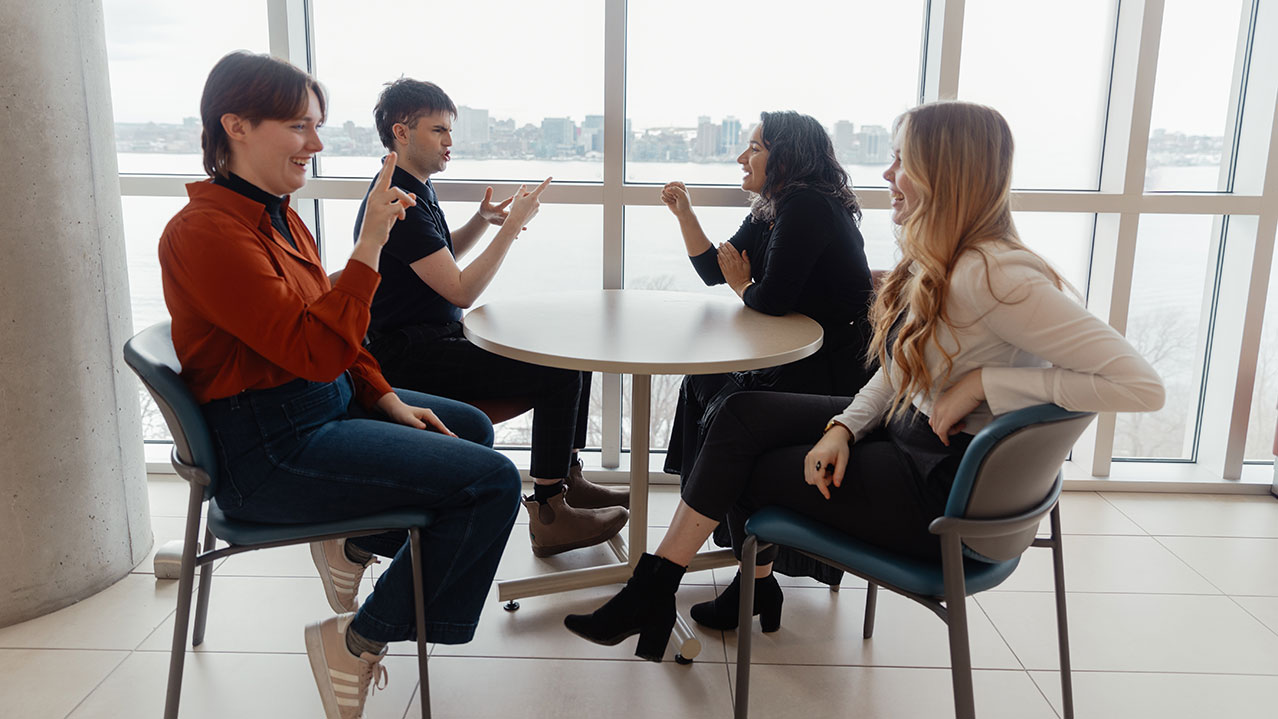 Four ASL students laugh while sitting in front of a window overlooking the Halifax Harbour signing to one another.