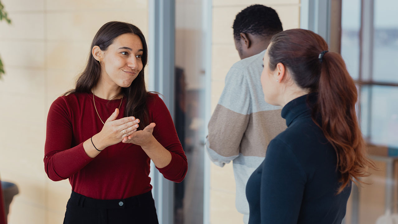 Two ASL students can be seen signing to one another outside their classroom while a third student attempts to enter the class