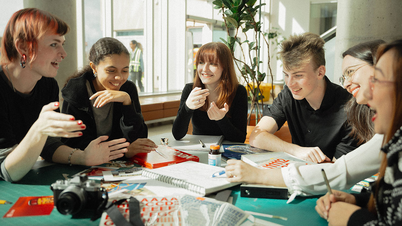 A group of students are seen sitting at a round table discussing a creative solution to their work.