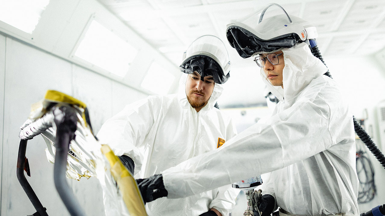 Two students in the auto body and collision repair program are seen in white suits, protective helmets and gloves working on an auto part together.