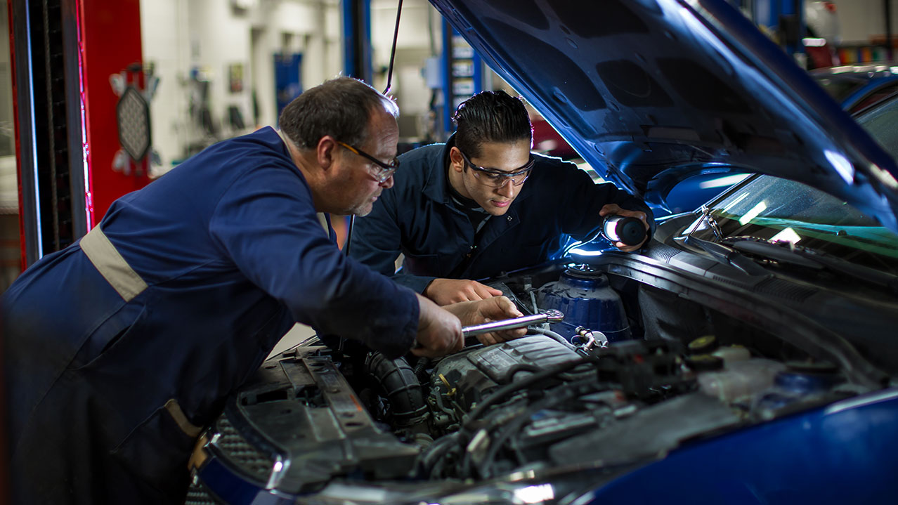 A student and instructor examine the engine of a car in an autobody shop.
