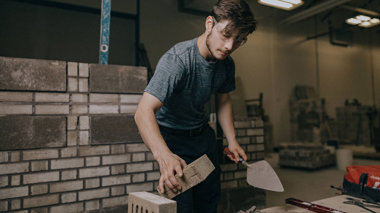 A student wearing safety glasses carefully places a brick in place at a work site.