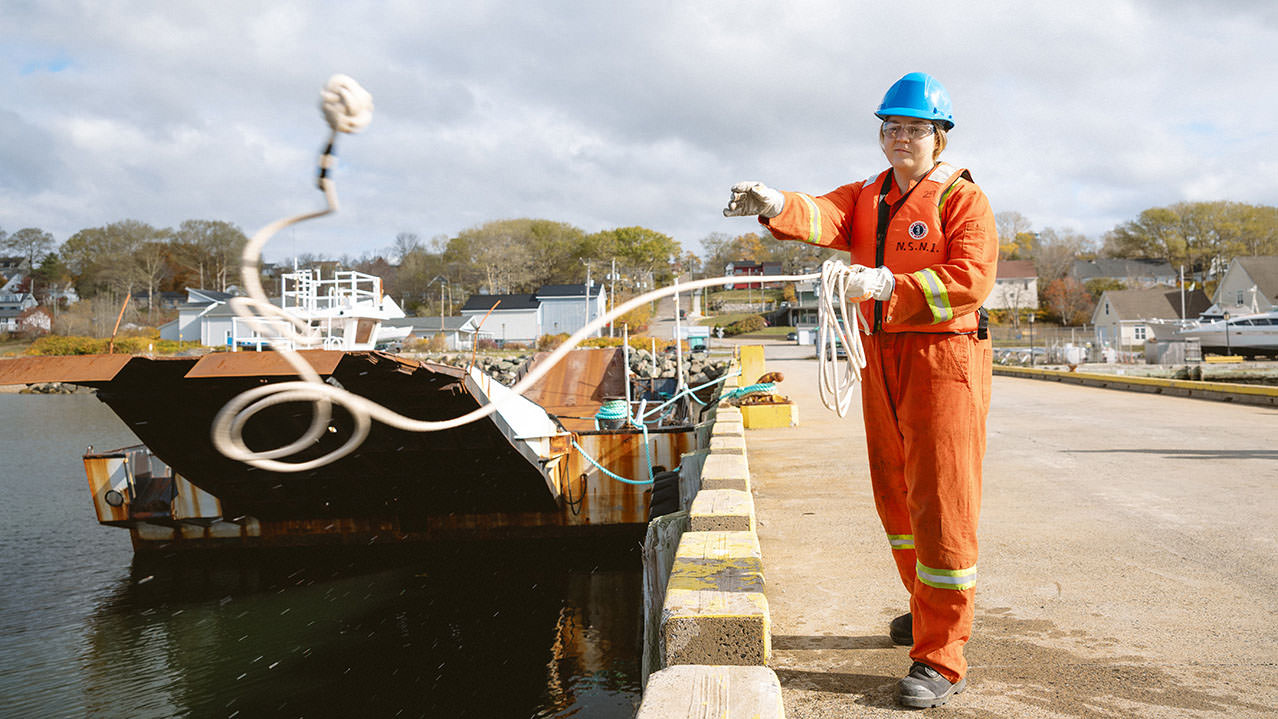 A student is seen standing on a dock in bright orange coveralls and blue hard hat, throwing a heavy rope out to a ship about to dock.