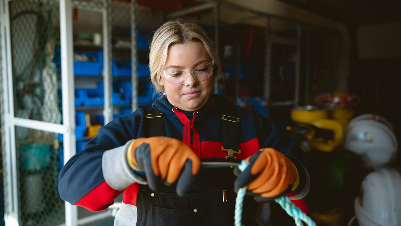 A female student in orange gloves and warm raincoat is seen working on a ship. 