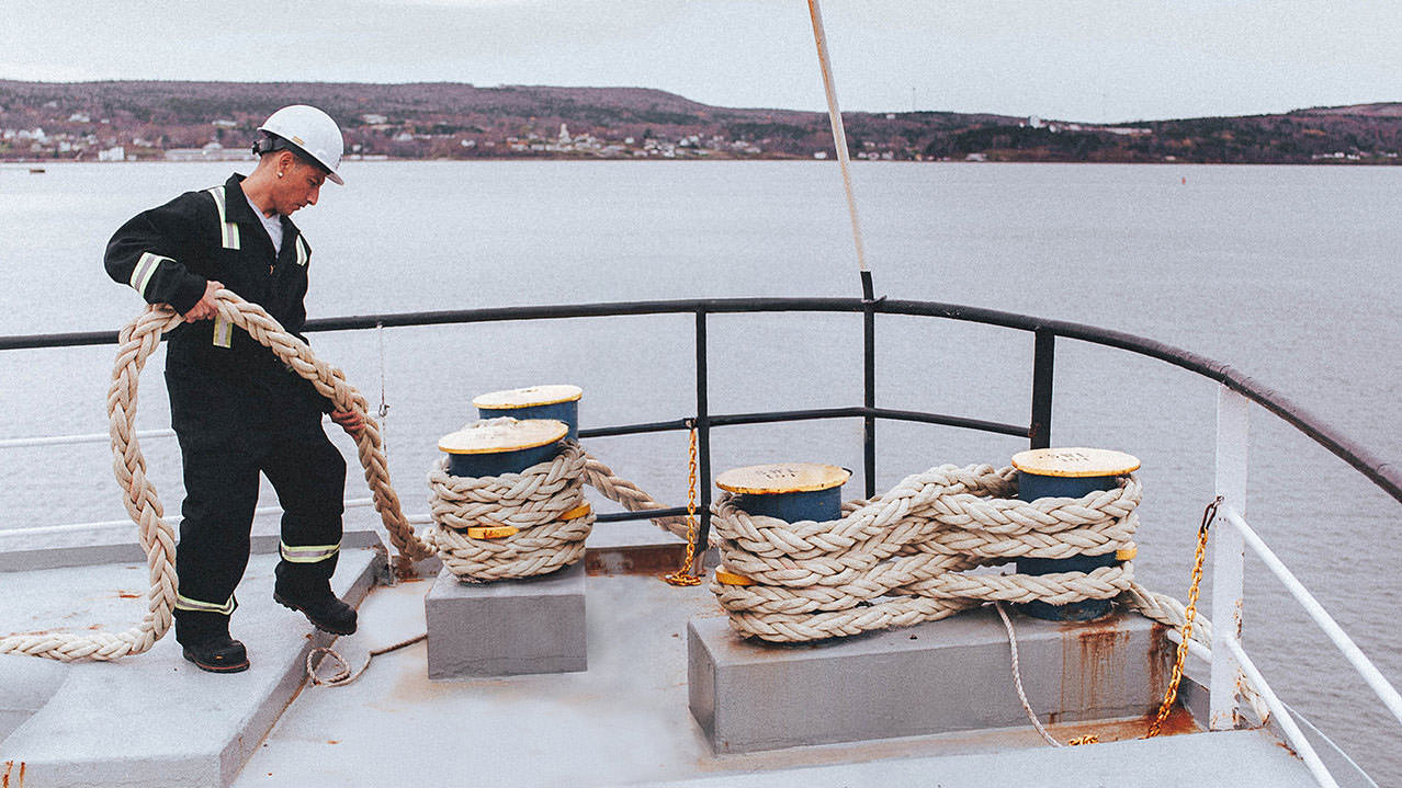 A student wearing coveralls and a hard hat is seen holding rope on a boat out at sea.