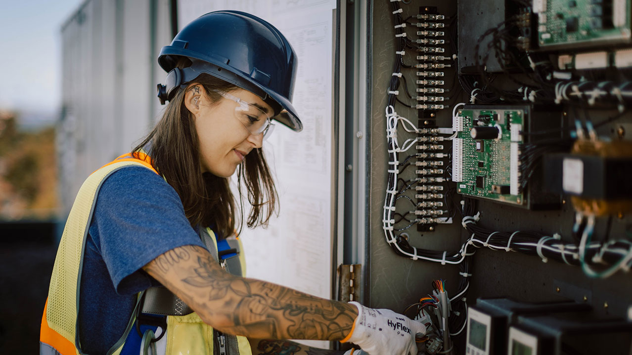 An HVAC professional wearing a safety hat, vest, glasses and gloves looks at wiring.
