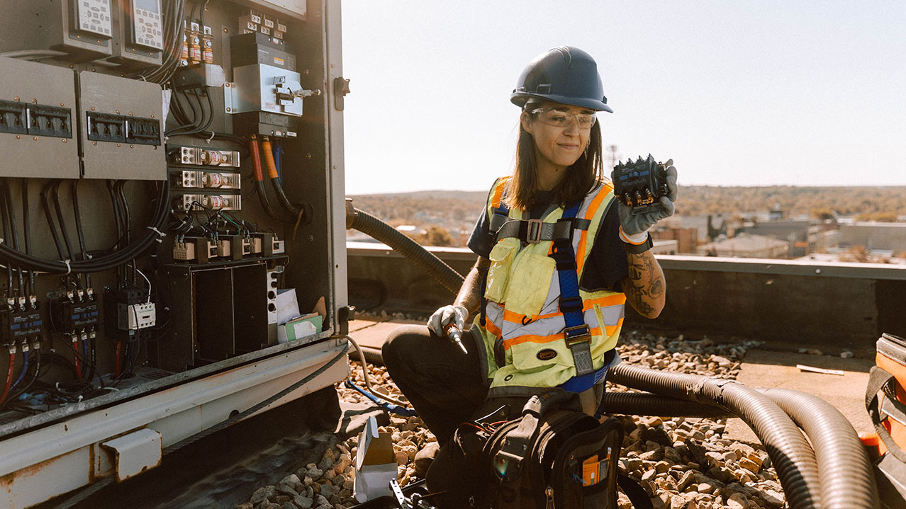 A woman in a safety vest and hard hat kneels looking at a piece of HVAC equipment.