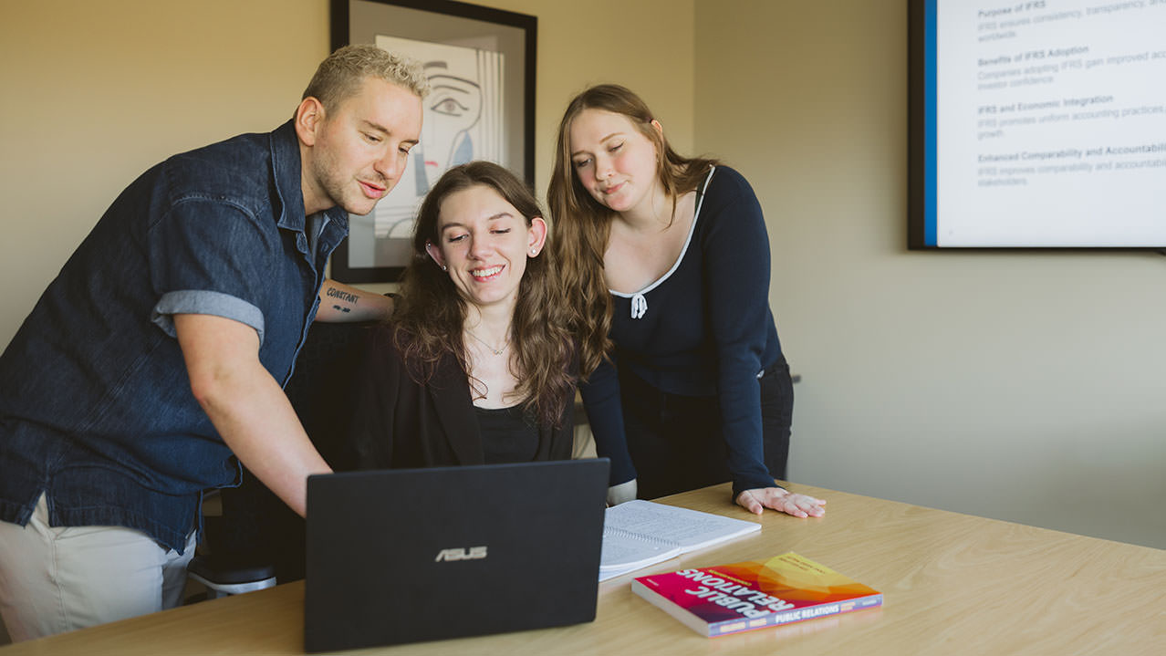 Three Business Administration students gather around a table and look at an open laptop together.