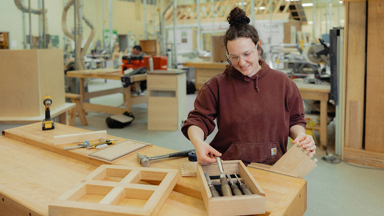 A female student in safety glasses is seen in a woodworking workshop choosing tools for her project.