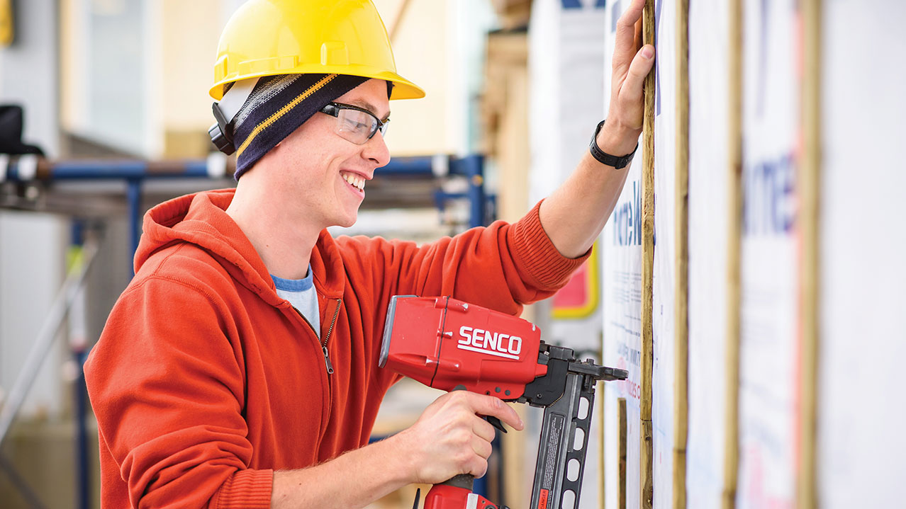 A carpentry student wearing a sweater, safety glasses and a hard hat uses a tool to construct a wooden frame for a wall.