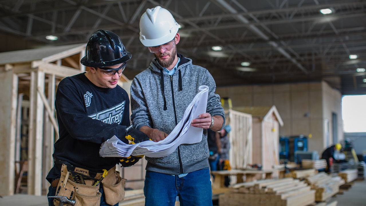 Two carpentry students wearing hard hats and safety glasses look at printed construction plans.